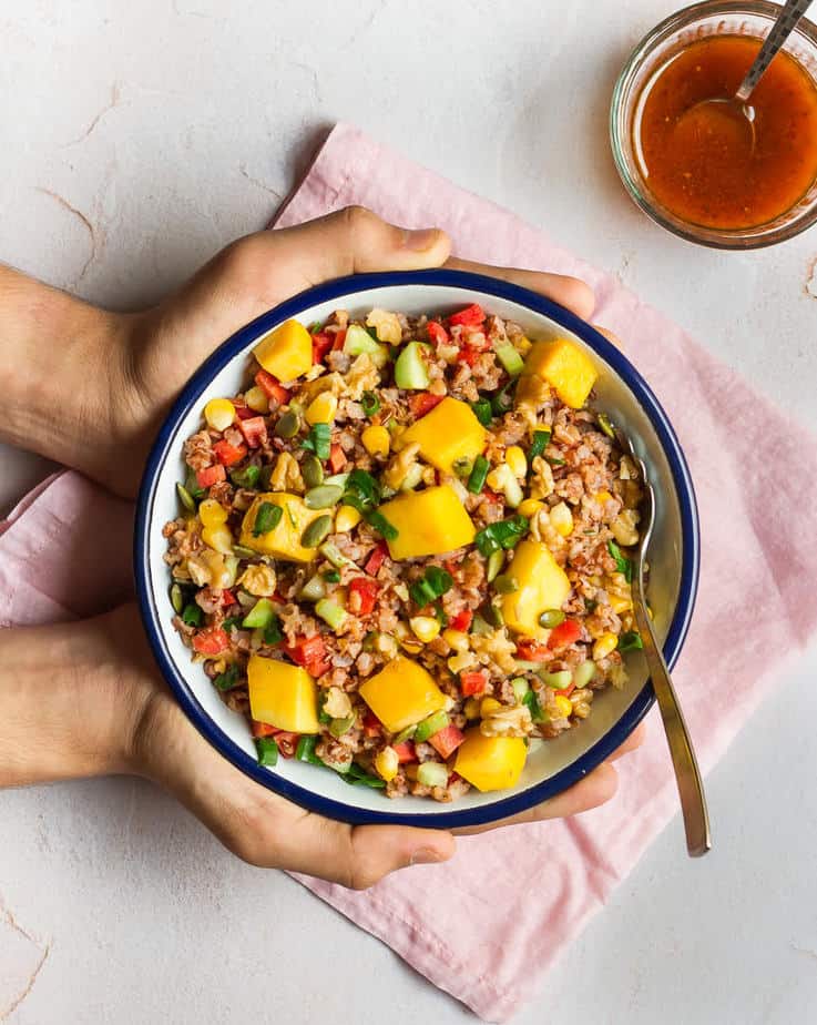 Hands holding a bowl of red rice and mango salad.