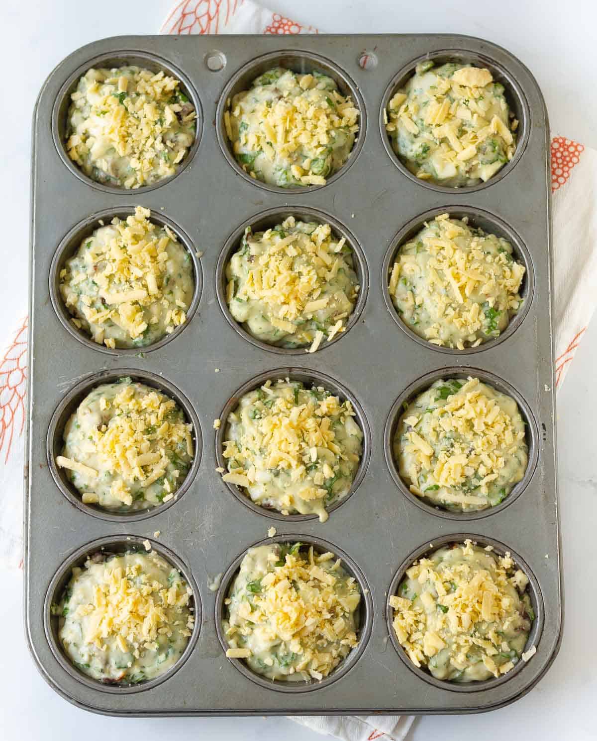 Muffin batter topped with shredded cheese in a muffin tray.