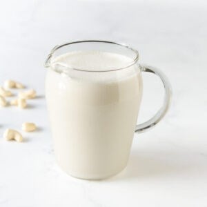 cashew milk in a glass pitcher with some cashews in the background.