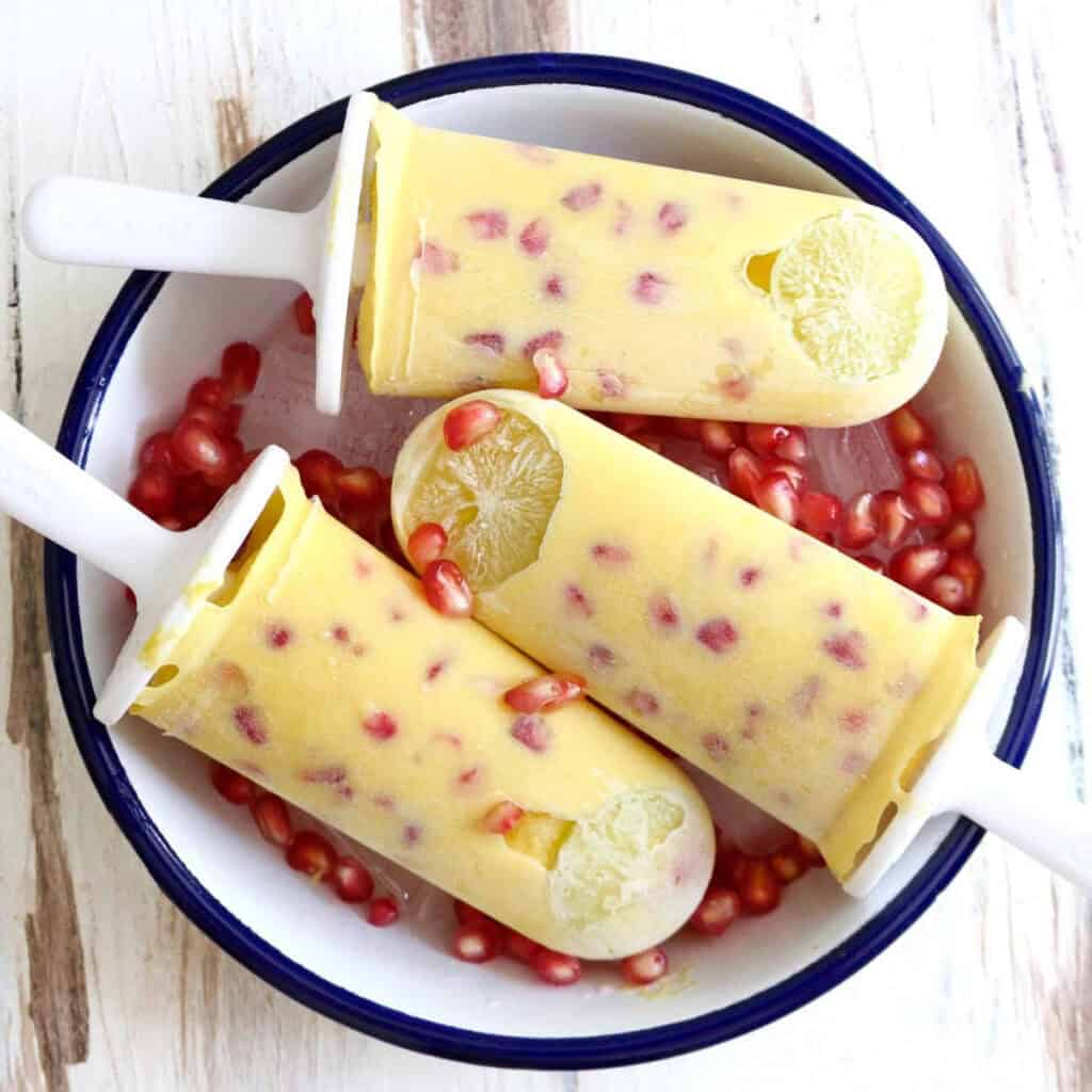 3 mango popsicles in a white bowl with pomegranates and ice.