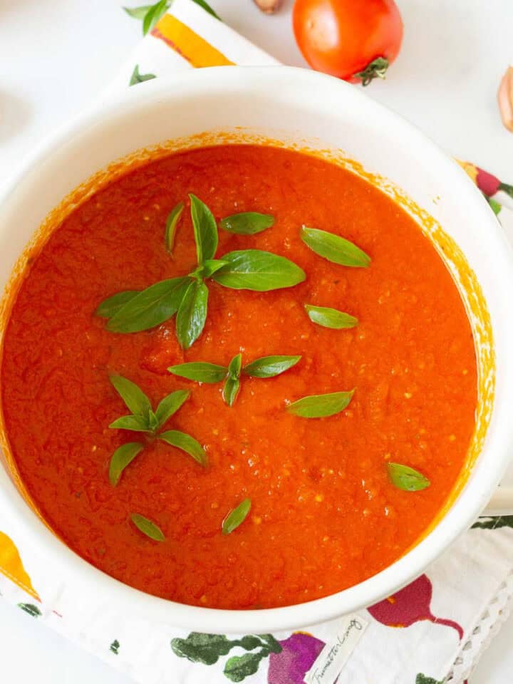 A white stockpot with tomato sauce topped with basil leaves with ingredients around.