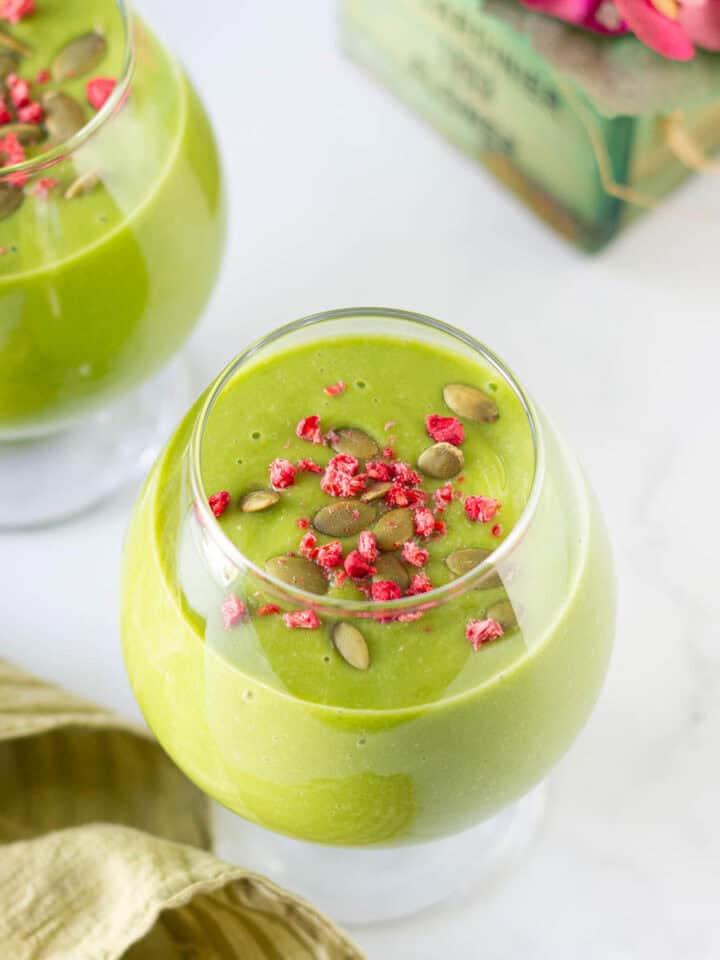 mango matcha smoothie topped with dried raspberries and pumpkin seeds in 2 glasses.