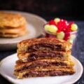A stack of half chocolate stuffed pancakes on a plate with fresh fruits around.