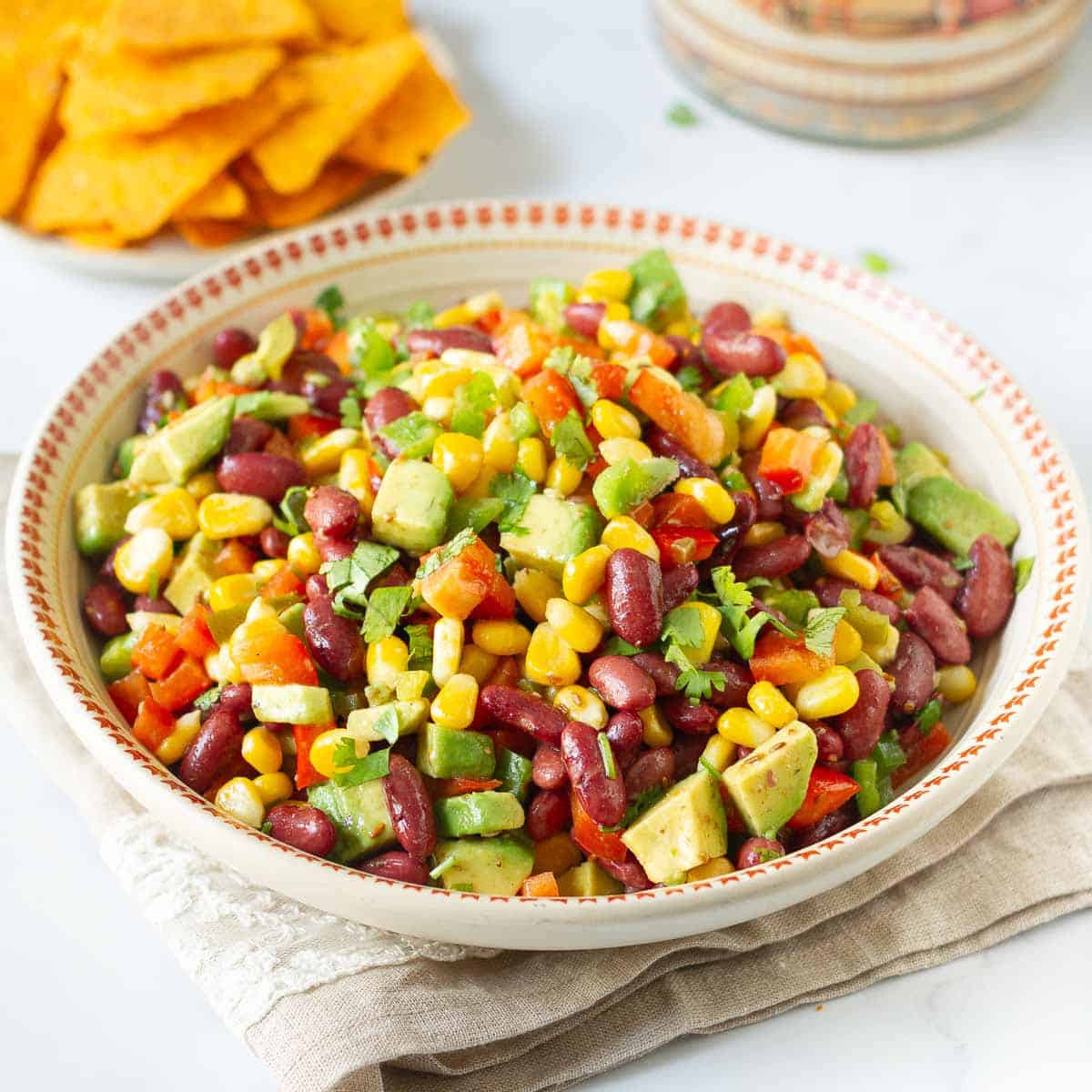 A bowl of bean avocado salad on a napkin with nachos in the background.