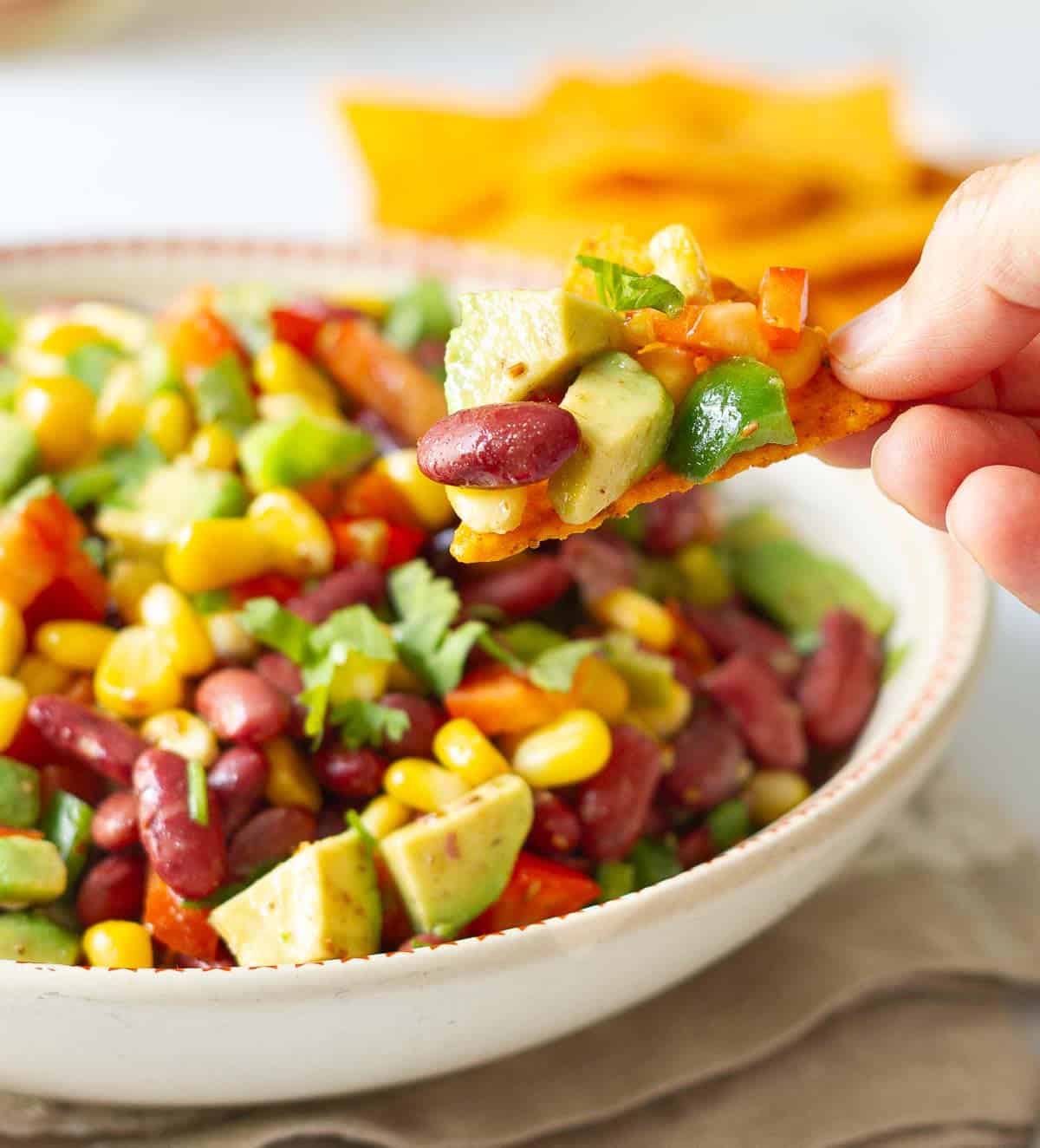 A hand holding a nacho with corn bean salad on top.
