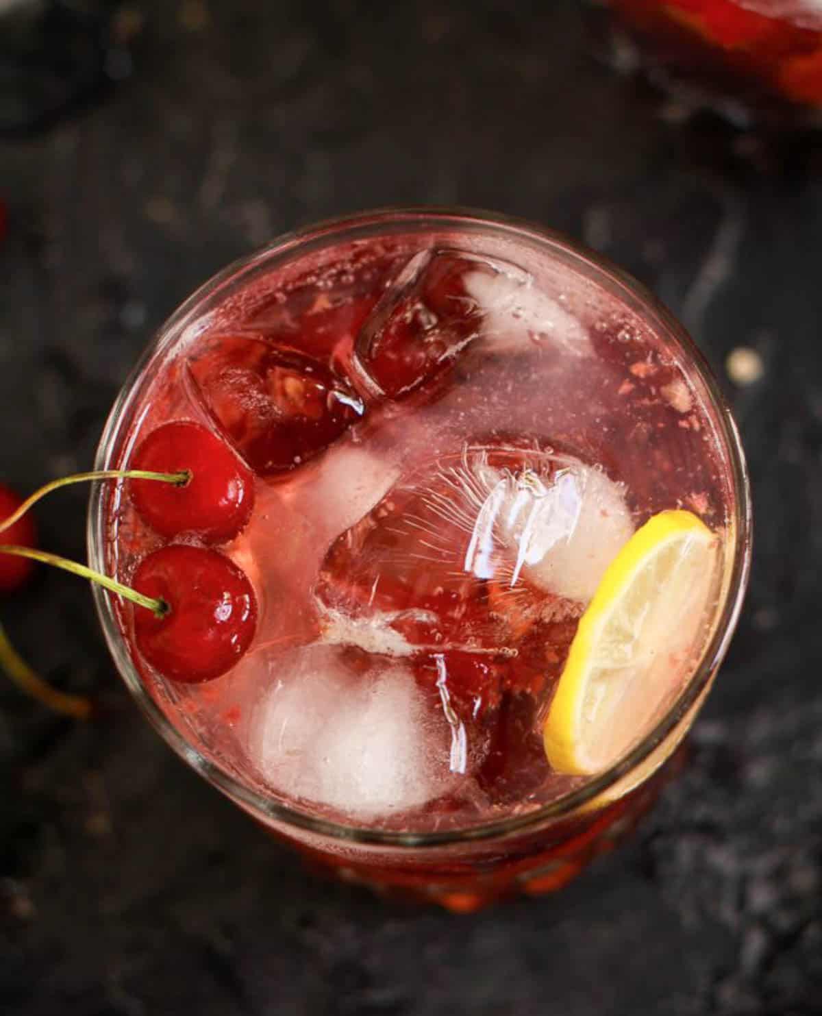 A glass of gin and tonic with cherries, ice, and lemon slice.
