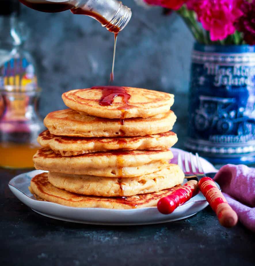 A stack of pancakes on a plate with a maple syrup getting poured on top.