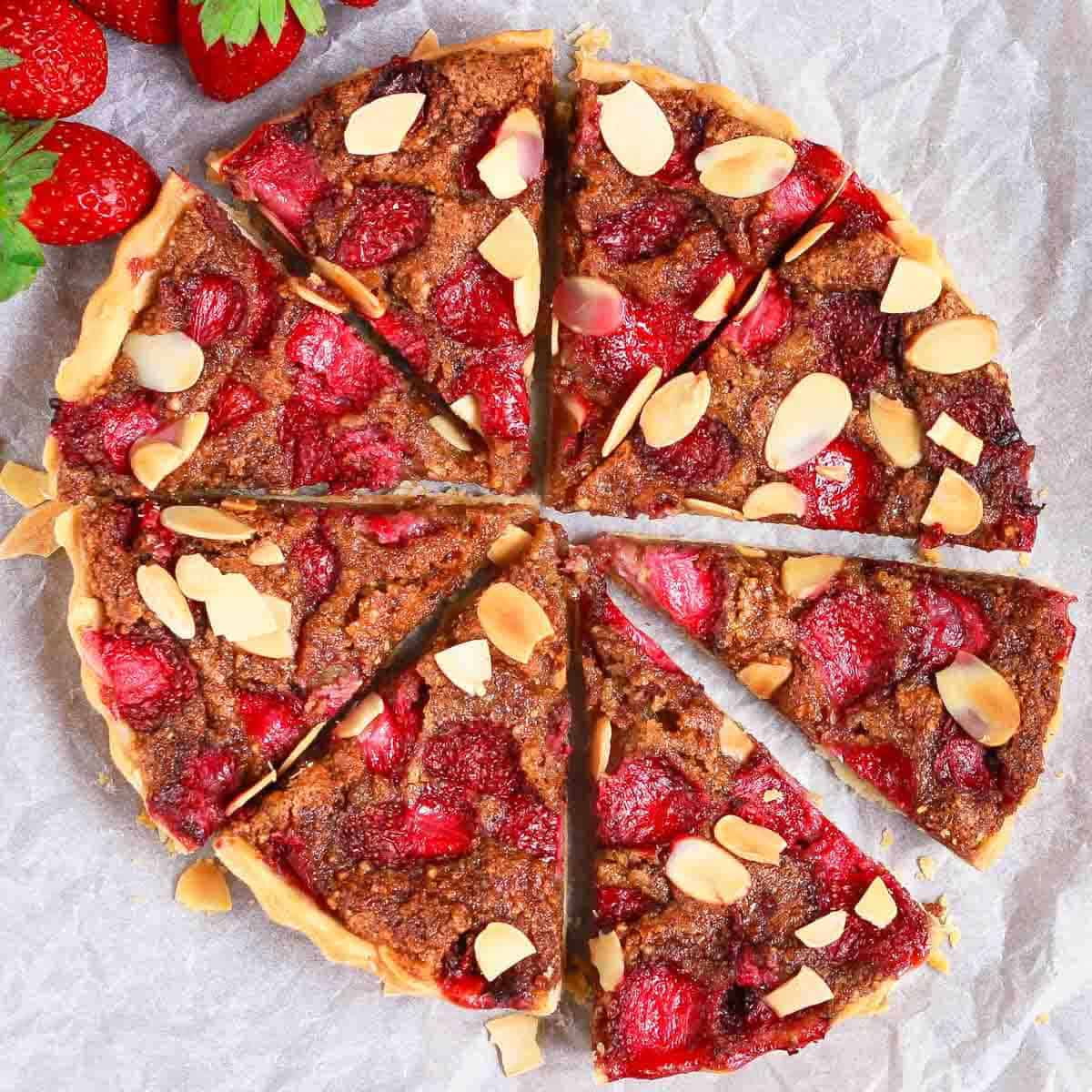 A sliced tart with almonds and strawberries on parchment paper.