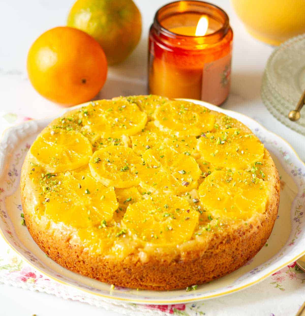 Orange upside down cake on a platter with a candle and oranges around.
