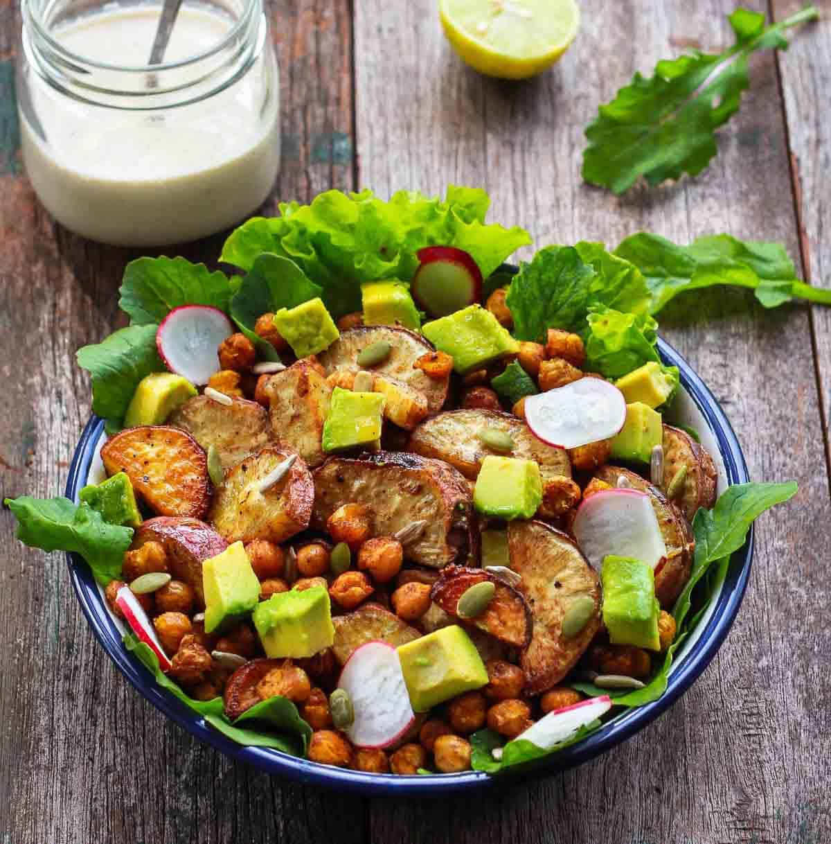 A bowl of salad with a jar of dressing and half a lemon on a wooden surface.