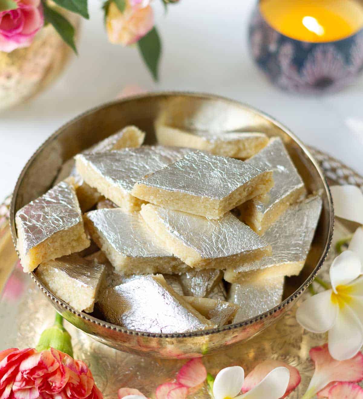Kaju burfi in a silver bowl with candle and flowers around.