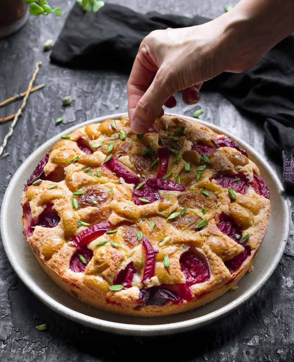A hand putting pistachios on a cake with plums and apricots on a platter.