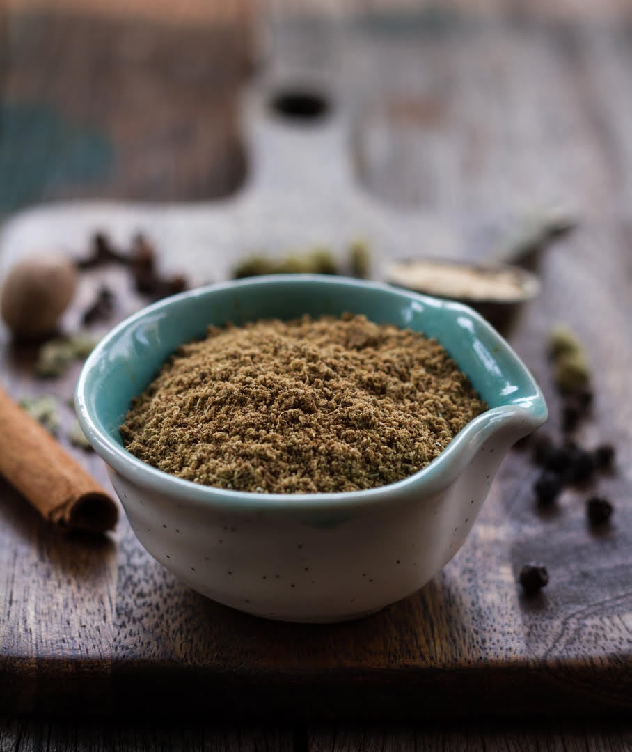 chai masala in a small bowl on a wooden board with ingredients in the background.