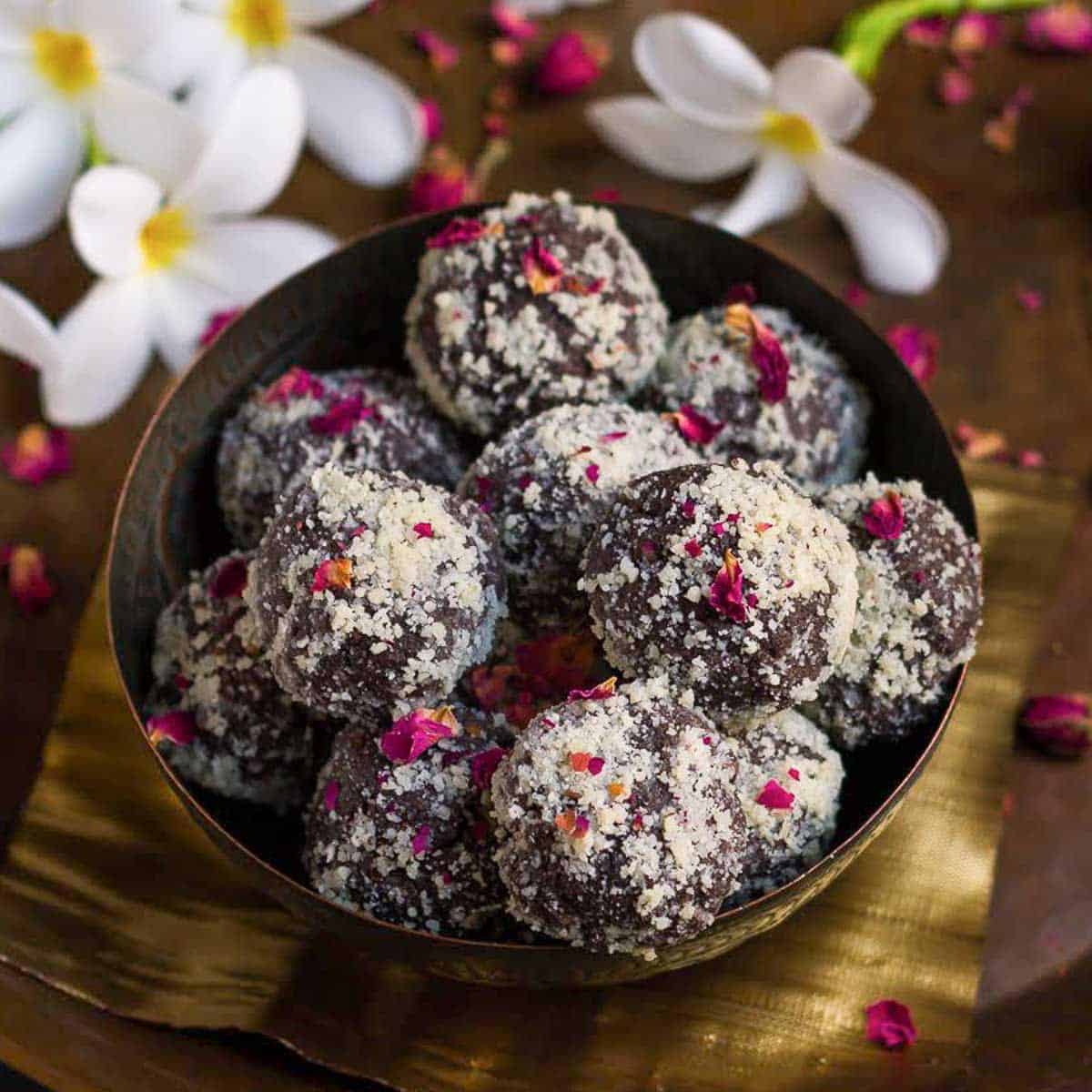 Chocolate pedas rolled in almond meal garnished with rose petals in a bowl with flowers around.