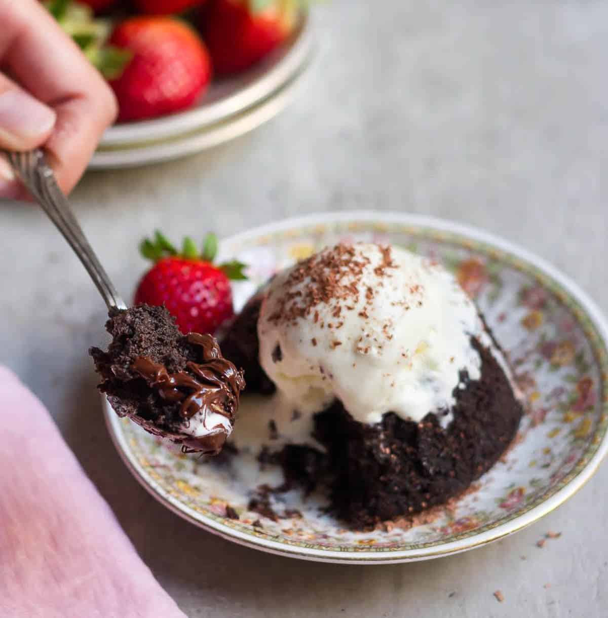 A hand holding a spoon of chocolate cake and a plate of cake with ice cream on top.