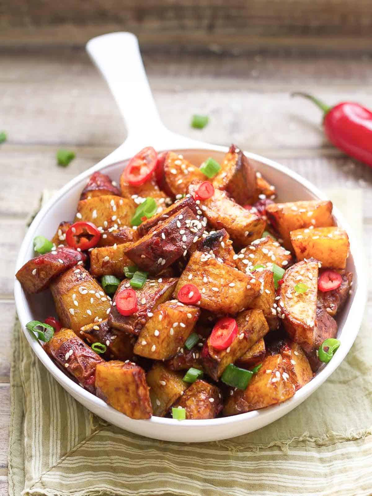spicy Sweet potatoes in a white pan with sesame seeds and green onions on top.