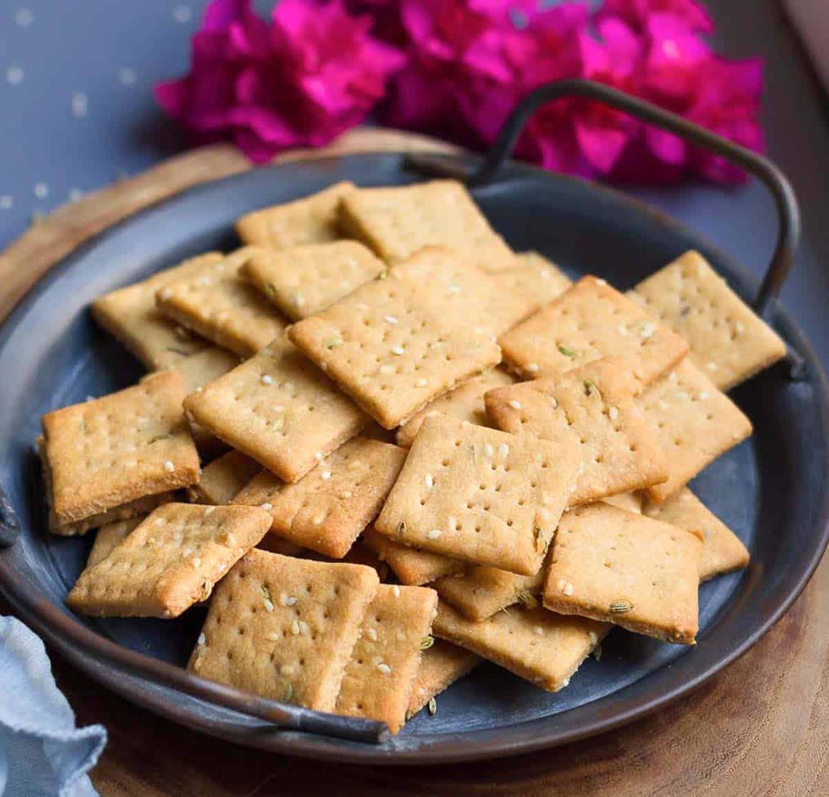 A batch of sweet mathri on a grey tray with flowers in the background.