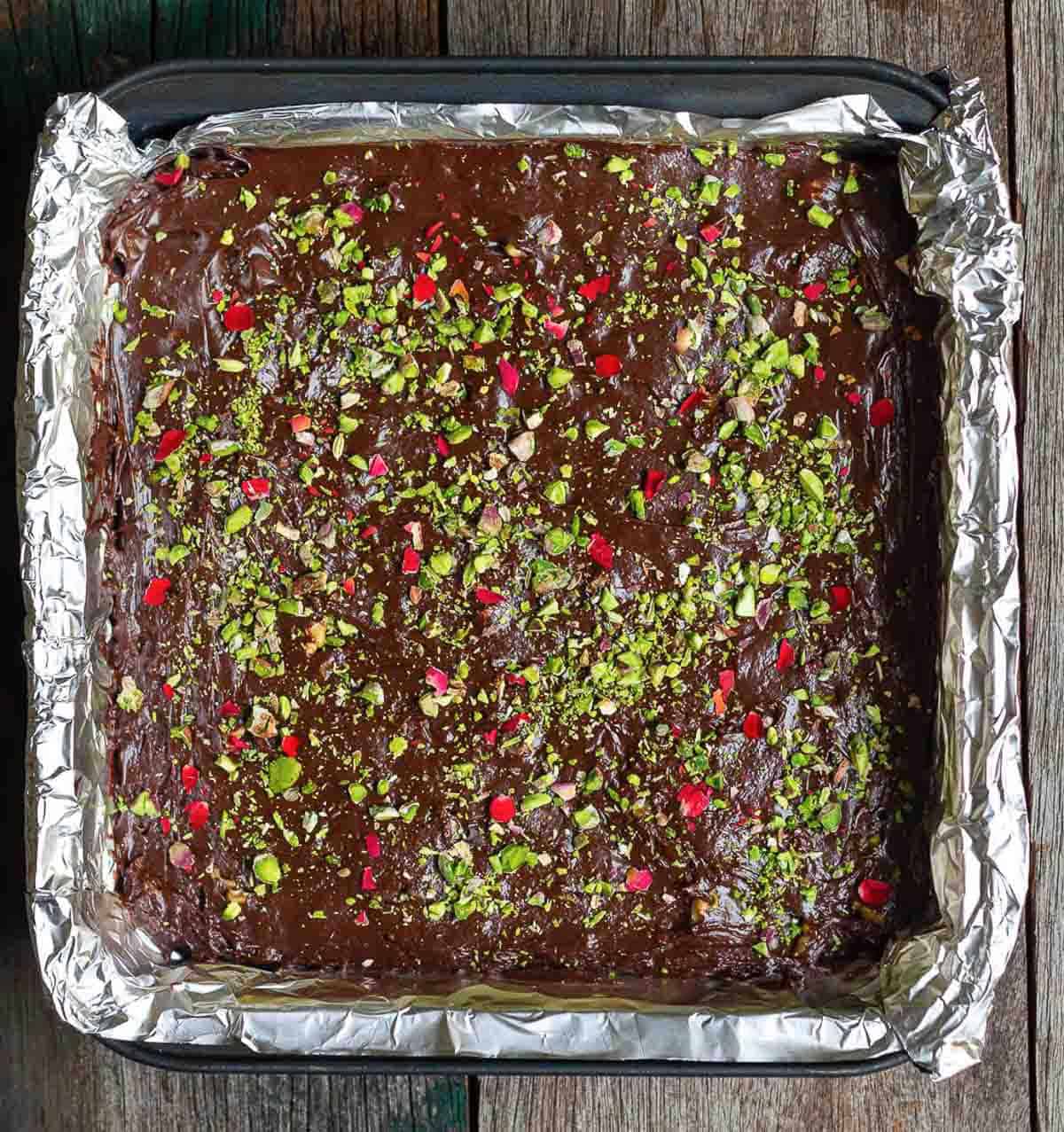 chocolate fudge topped with pistachios, walnuts, rose petals in a square pan lined with foil.