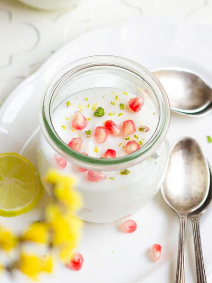 A jar of panna cotta garnished with pomegranate arils kept on a white platter with spoons.