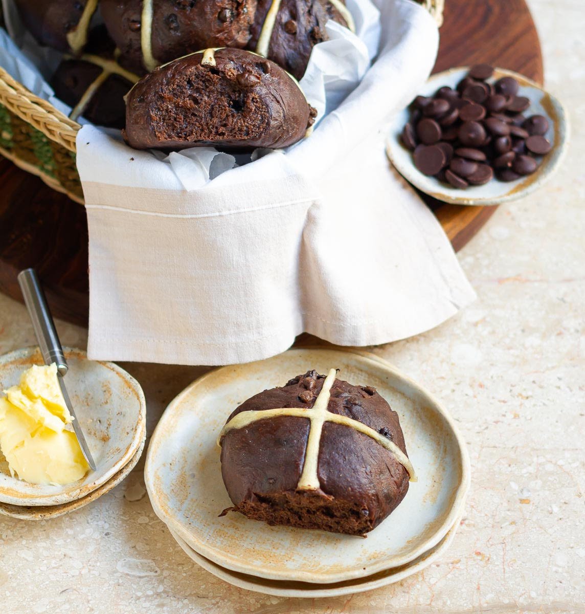 Hot cross buns in a basket and a small plate, surrounded by a small plate of butter and chocolate chips