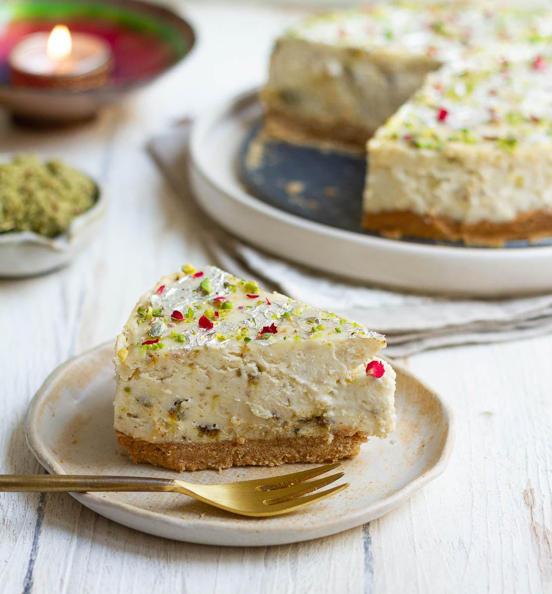 A slice of cheesecake , festive candle, thandai masala and cheesecake on a platter