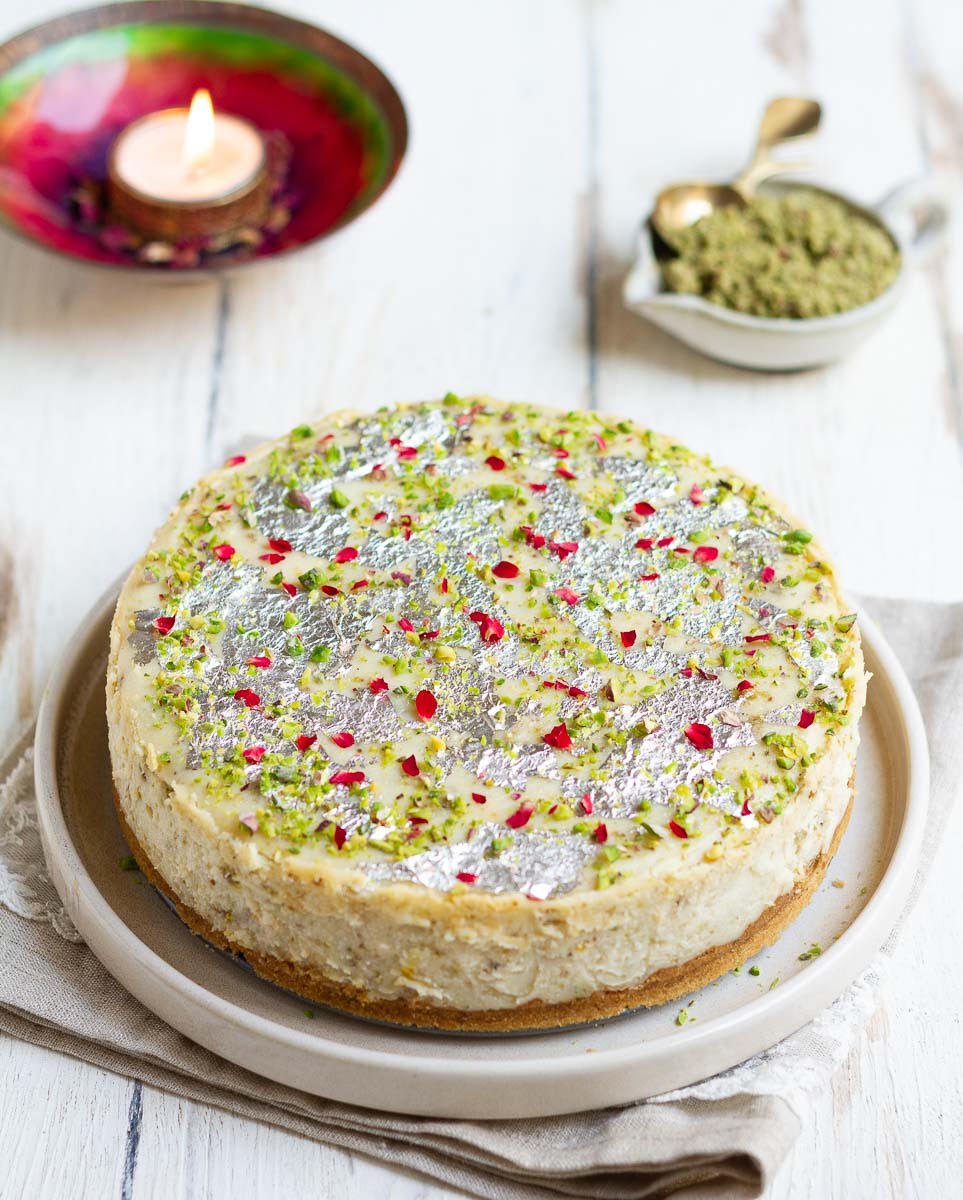 Decorated cheesecake on a platter with a festive candle and thandai masala in a small bowl