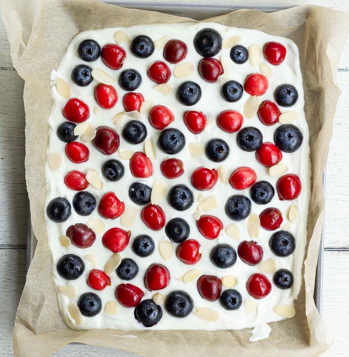 Yogurt spread out on a parchment lined tray and blueberries, cherries and almond flakes.