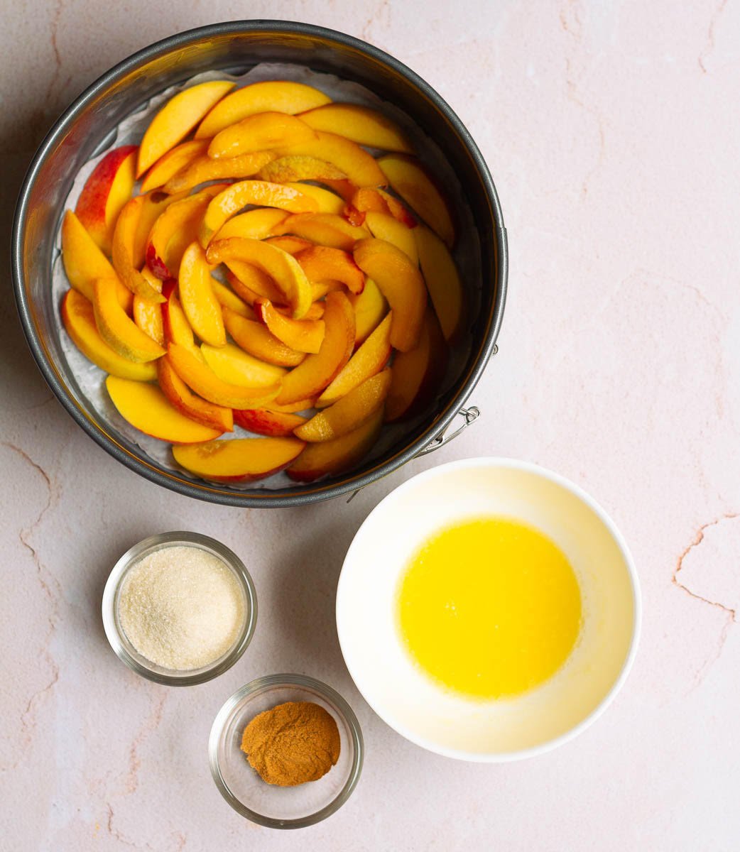 Sliced peaches lining the bottom of cake tin and topping of sugar, butter, cinnamon on the side