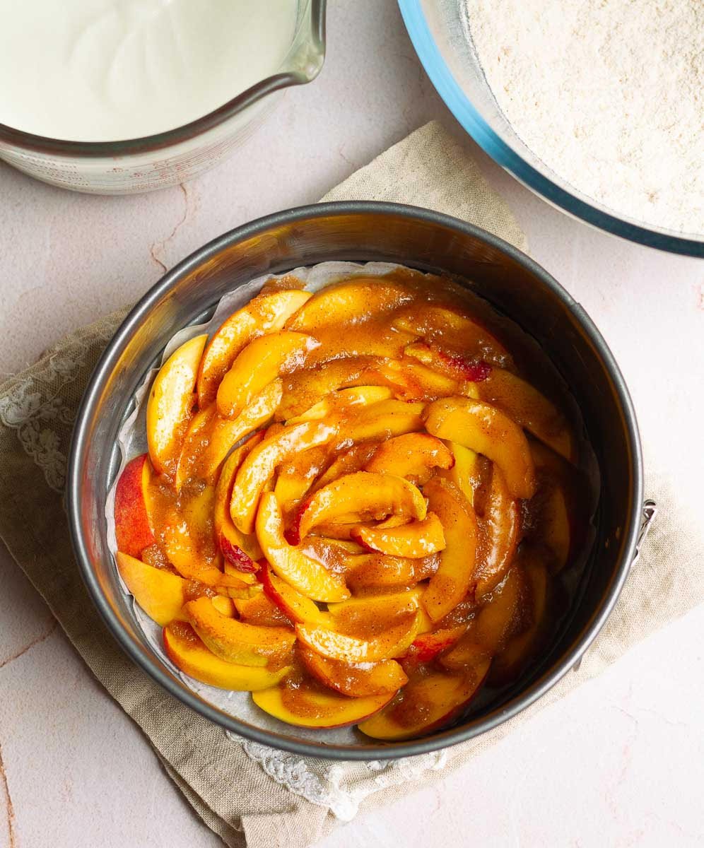 Dry and wet ingredients next to the cake tin with sliced peaches topped with cinnamon butter