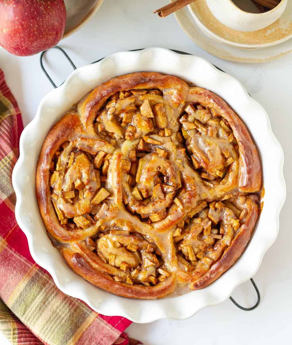 Apple Pie Cinnamon Rolls in a round pie dish on a trivet with ingredients in the background.