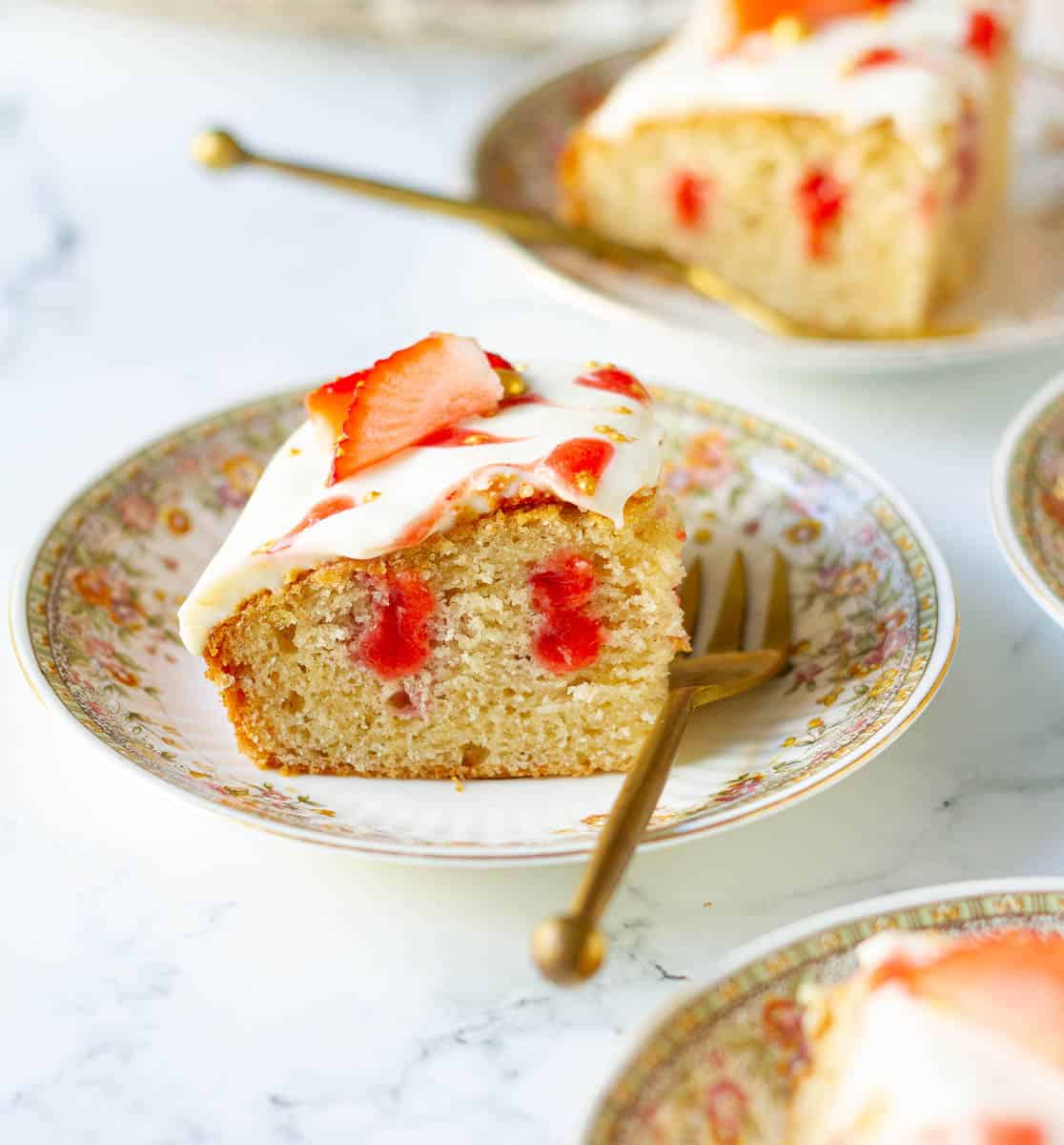 A square of strawberry cake on a small plate with a golden fork.