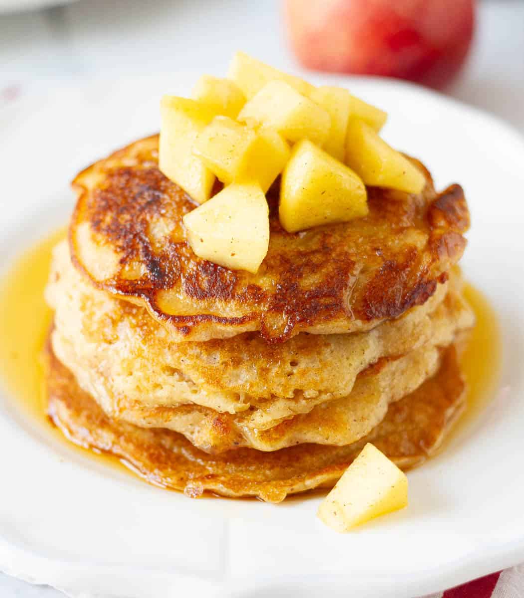 stack of apple pancakes topped with maple syrup and apples on a white plate.