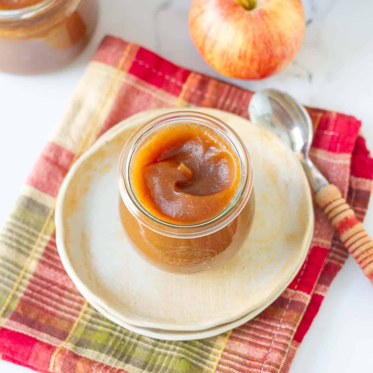 Stovetop apple butter in a glass jar with a spoon on a plate with apple and apple butter in the background.
