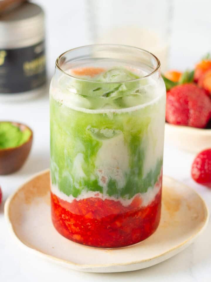 strawberry matcha latte in a glass on a small plate with ingredients in the background.