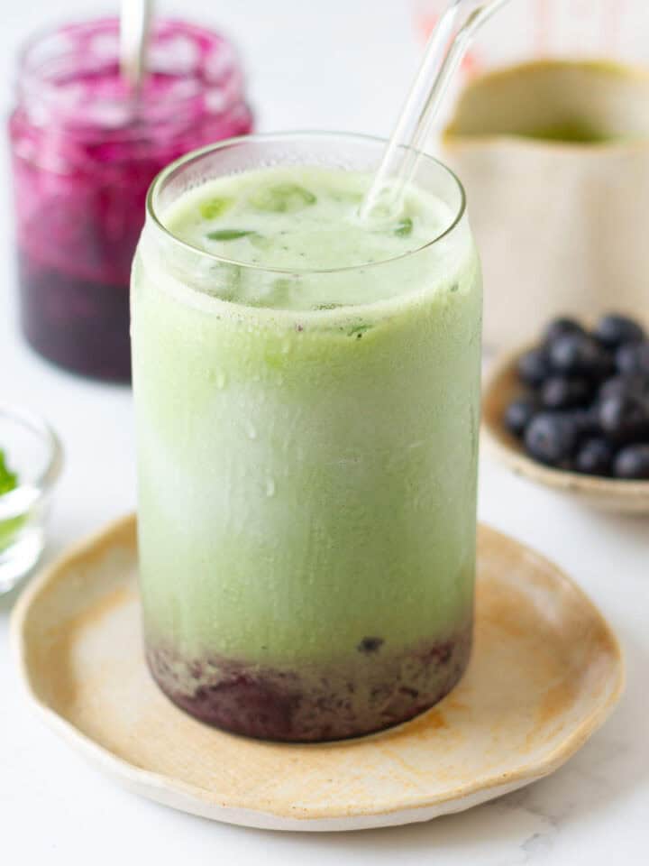 Blueberry matcha latte in a glass with blueberry syrup and blueberries in the background.