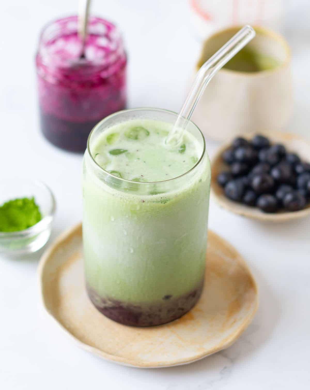 iced blueberry matcha latte with a glass straw with ingredients in the background.
