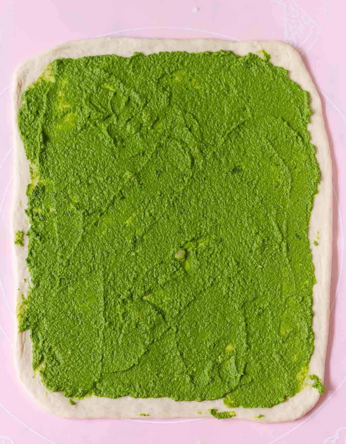 Rolled out dough with pesto on top on a pink silicone mat.
