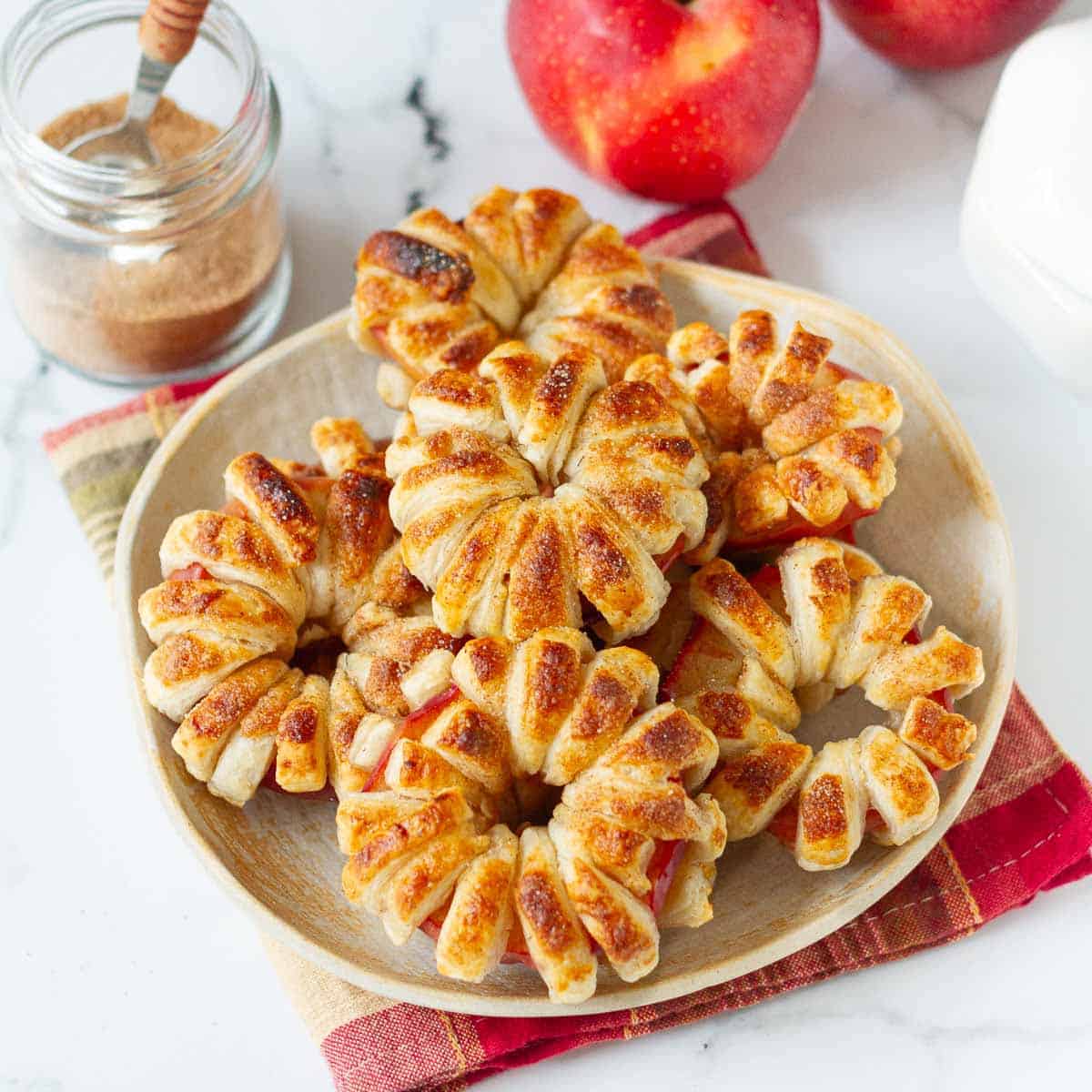 Golden puff pastry apple donuts on a plate with apples and cinnamon powder in the background.