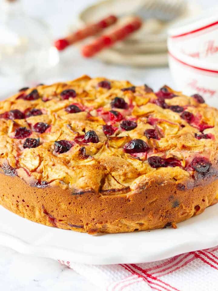 Apple cranberry cake on a white plate with plates and cups in the background.