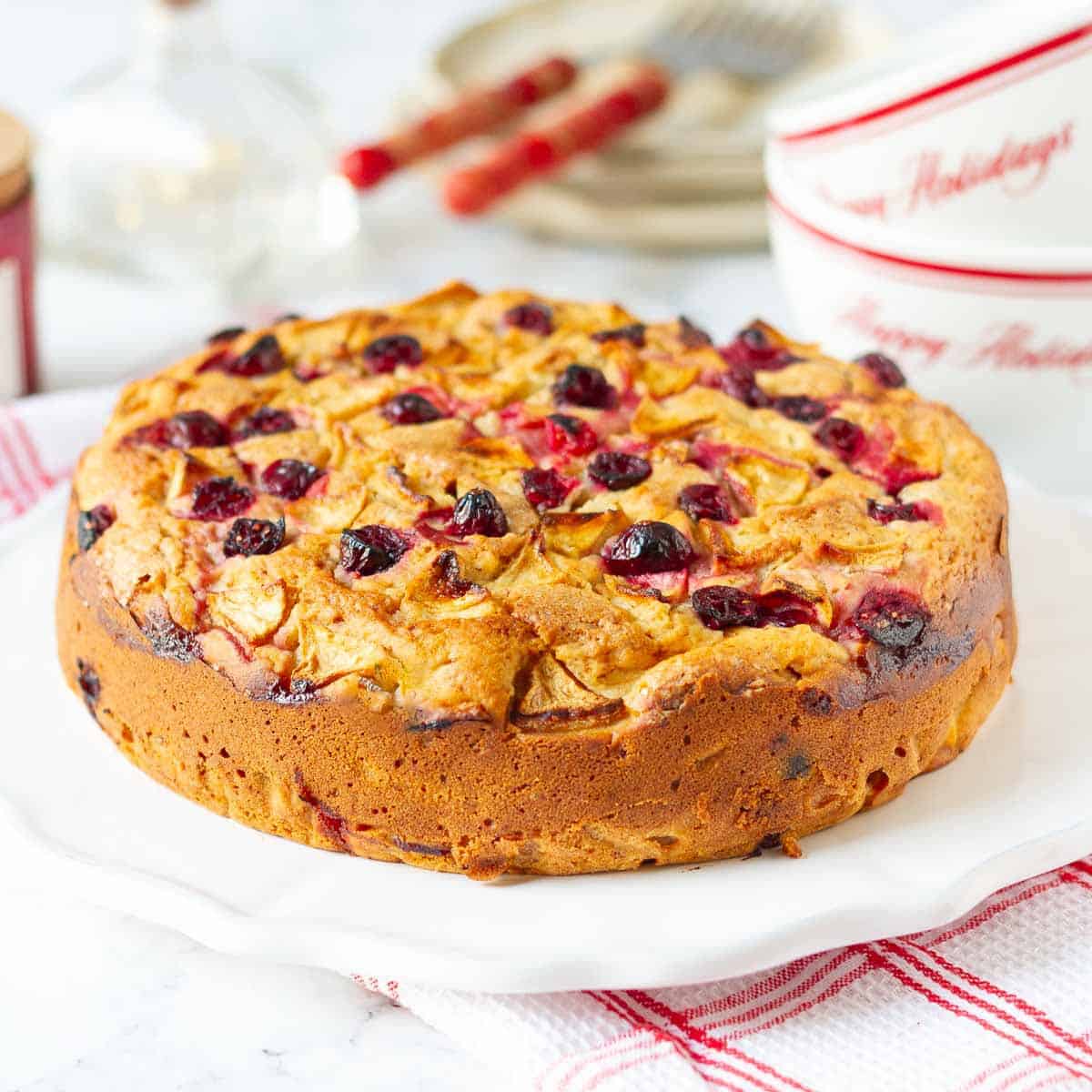 Apple cranberry cake on a white plate with plates and cups in the background.