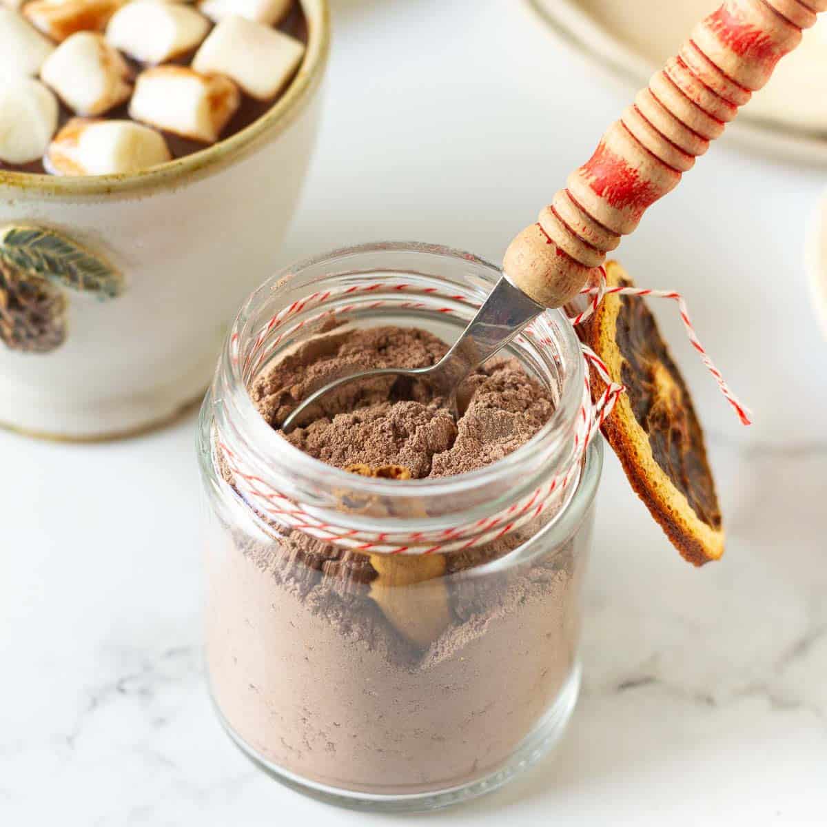 A jar of cocoa mix with a spoon and a cup of hot cocoa with marshmallows.