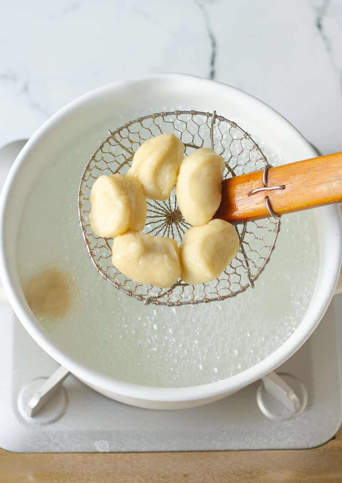 Pieces of dough on a spider strainer on top of a pot of boiling water.