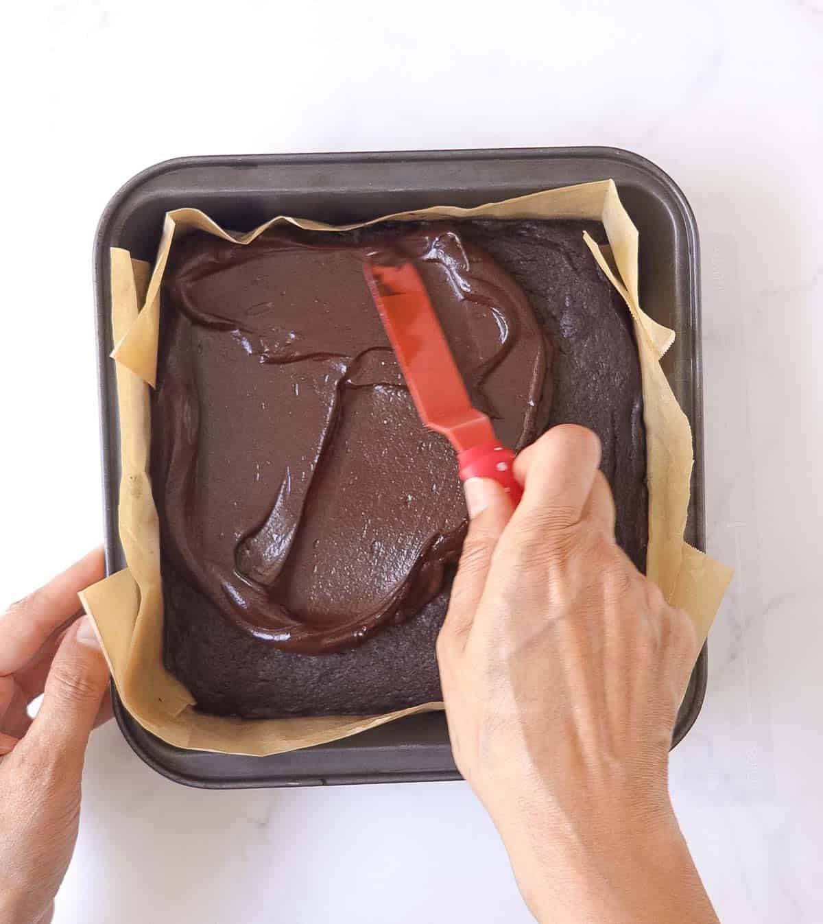 A hand spreading frosting on a brownie with a red palette knife.