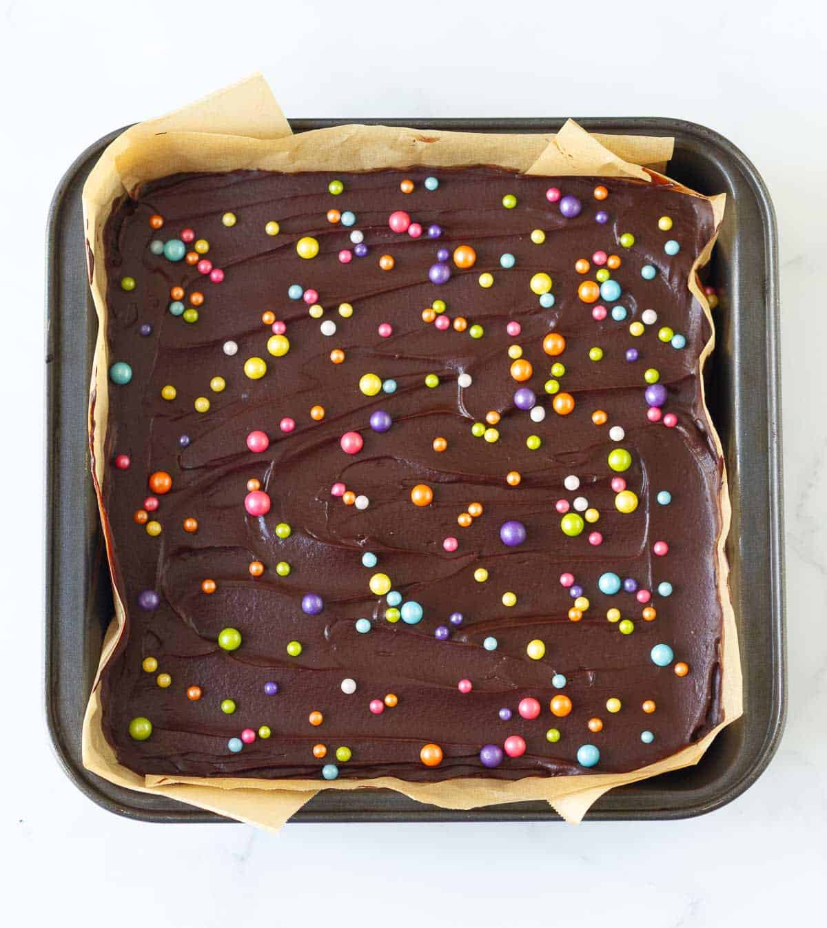 Brownie in a parchment-lined square pan with chocolate frosting and rainbow sprinkles.