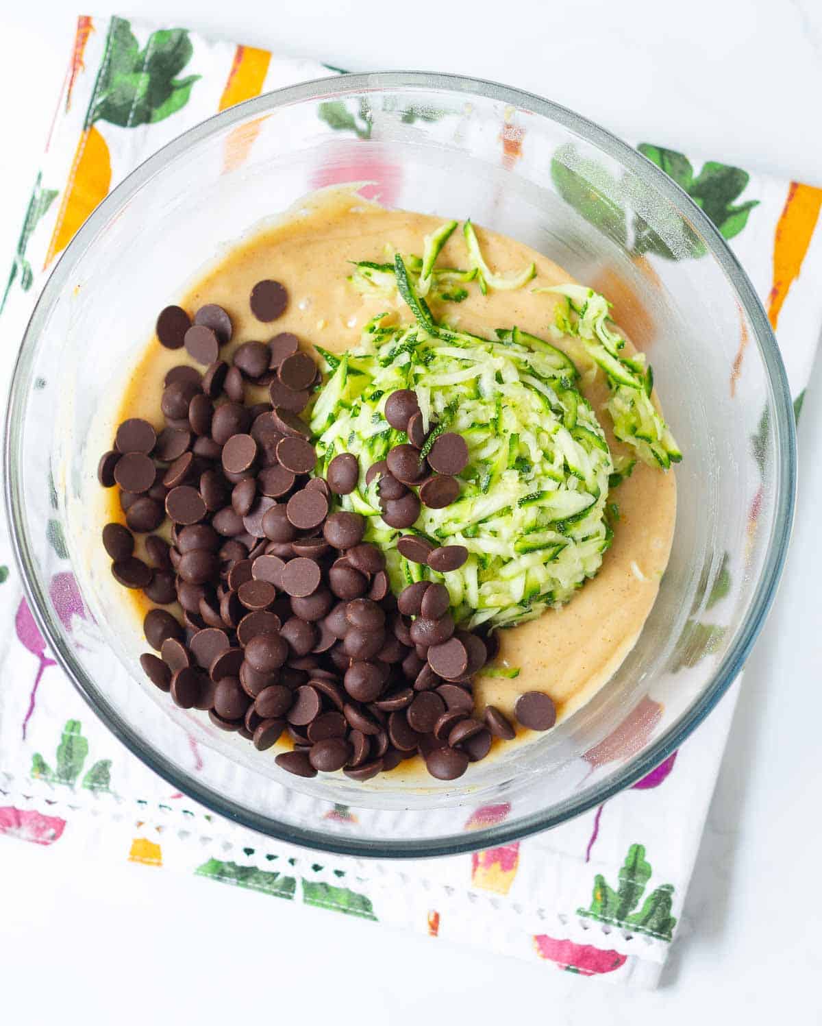 Shredded zucchini and chocolate chips on top of batter in a mixing bowl.