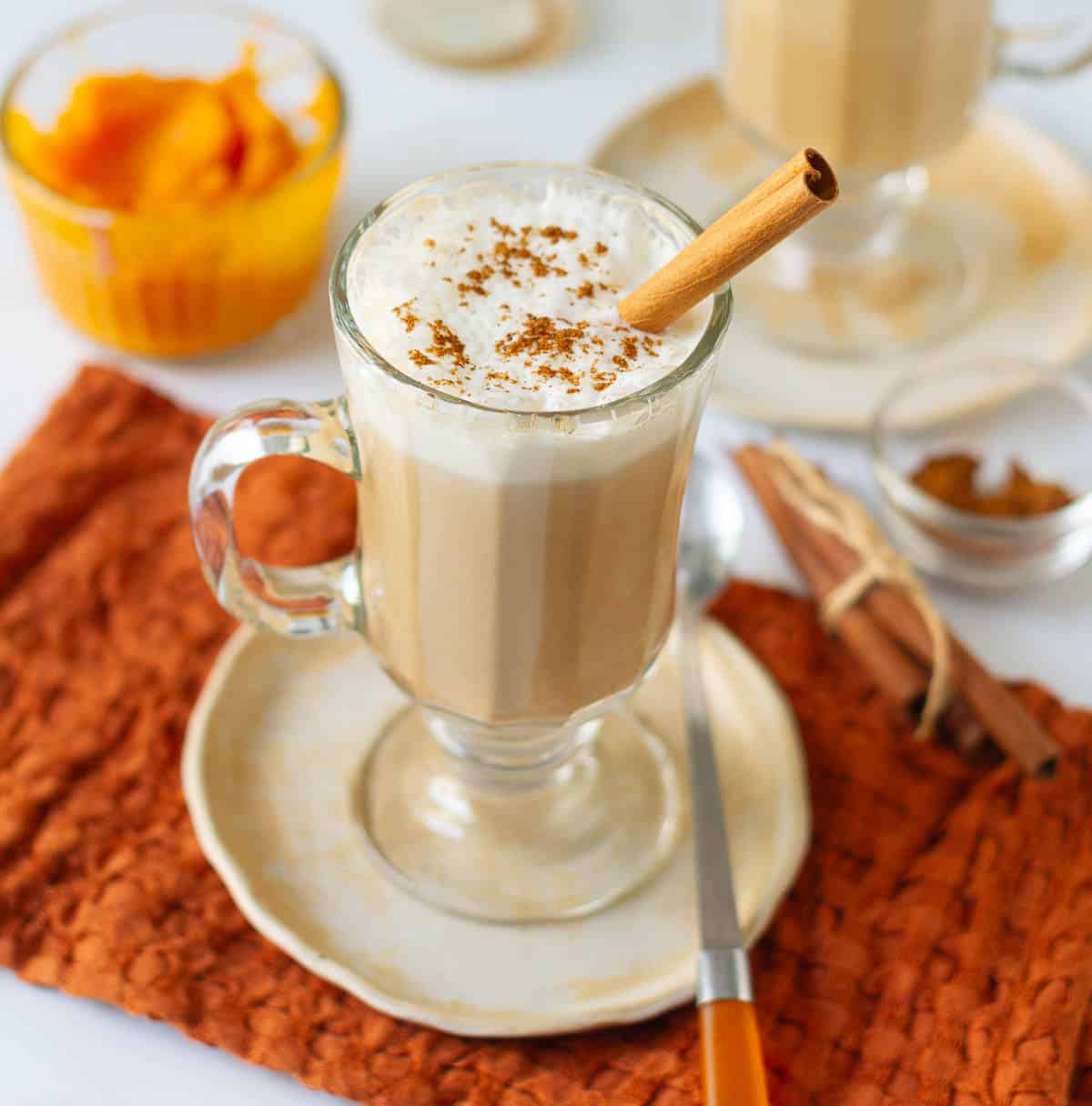 Pumpkin spice latte in a glass mug topped with cream and cinnamon stick on a small plate.